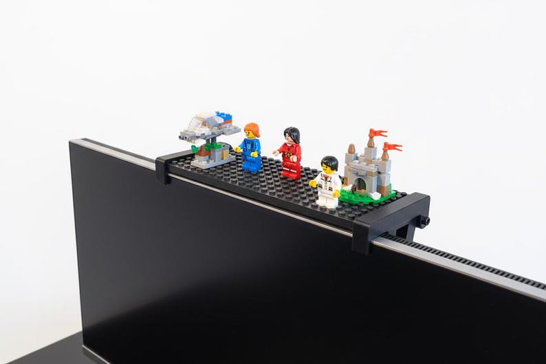 May include: A black monitor shelf with a black studded surface holds several Lego structures and minifigures. The Lego creations include a spaceship, a castle, and three minifigures in blue, red, and white outfits.