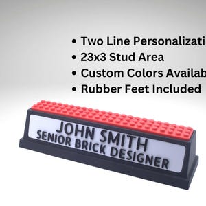 May include: A personalized nameplate with a black base, a white name area, and a red top. The nameplate reads "JOHN SMITH, SENIOR BRICK DESIGNER". Features include two-line personalization, a 23x3 stud area, custom colors, and rubber feet.