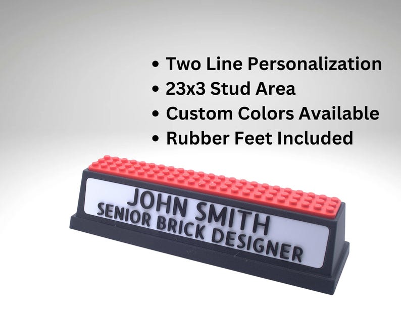 May include: A personalized nameplate with a black base, a white name area, and a red top. The nameplate reads "JOHN SMITH, SENIOR BRICK DESIGNER". Features include two-line personalization, a 23x3 stud area, custom colors, and rubber feet.