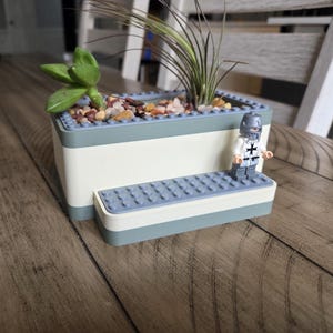 May include: A rectangular planter made from building blocks, with a light blue and white color scheme. The planter contains small rocks, a succulent, and a tall, thin plant. A toy knight stands on a small platform attached to the planter.