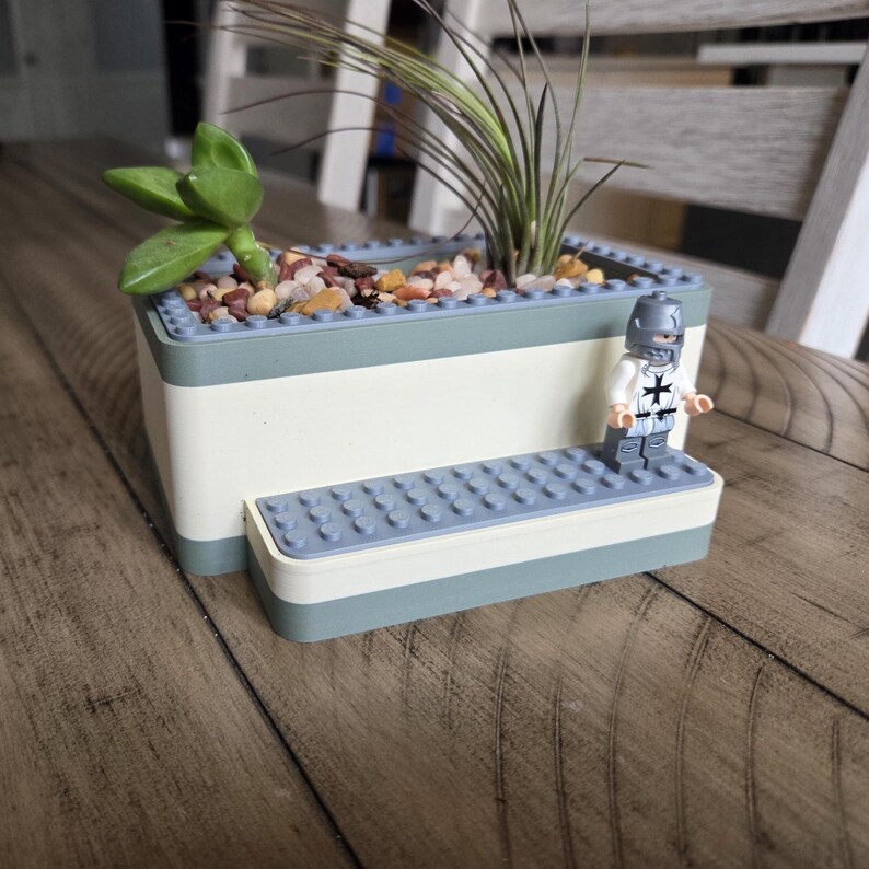 May include: A rectangular planter made from building blocks, with a light blue and white color scheme. The planter contains small rocks, a succulent, and a tall, thin plant. A toy knight stands on a small platform attached to the planter.
