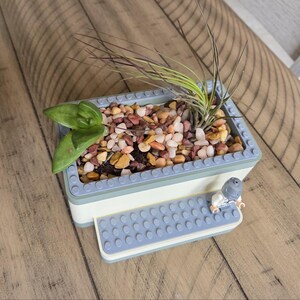 May include: A rectangular planter made from light blue and white blocks, filled with small colorful stones and greenery. A small Lego figure sits on a matching light blue platform attached to the planter.