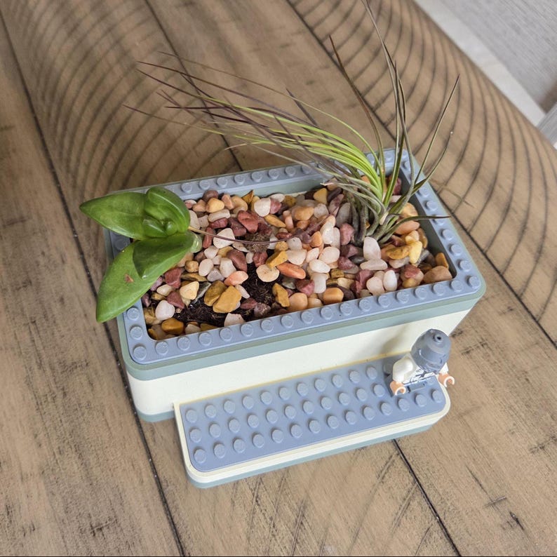 May include: A rectangular planter made from light blue and white blocks, filled with small colorful stones and greenery. A small Lego figure sits on a matching light blue platform attached to the planter.