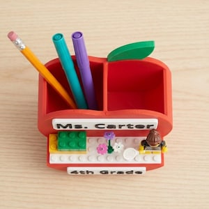 May include: A red apple-shaped desk organizer with two compartments containing a yellow pencil, a teal marker, and a purple marker. The front features a white sign reading Ms. Carter, a small building block display with flowers, and a small figure.