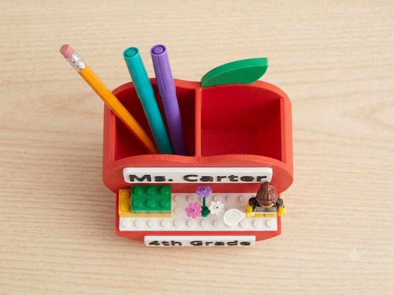 May include: A red apple-shaped desk organizer with two compartments containing a yellow pencil, a teal marker, and a purple marker. The front features a white sign reading Ms. Carter, a small building block display with flowers, and a small figure.