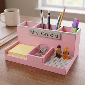 May include: A light pink desk organiser with three upper compartments for pens and markers, a lower section for sticky notes, a small tray for paper clips, and a grey building block base featuring two small plastic figures with yellow heads.