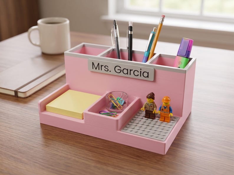May include: A light pink desk organiser with three upper compartments for pens and markers, a lower section for sticky notes, a small tray for paper clips, and a grey building block base featuring two small plastic figures with yellow heads.