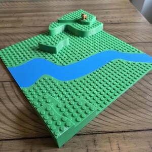 May include: A green building block baseplate with a blue river and raised green terrain. A small brown animal figurine is on the highest point. The baseplate is on a wooden surface.