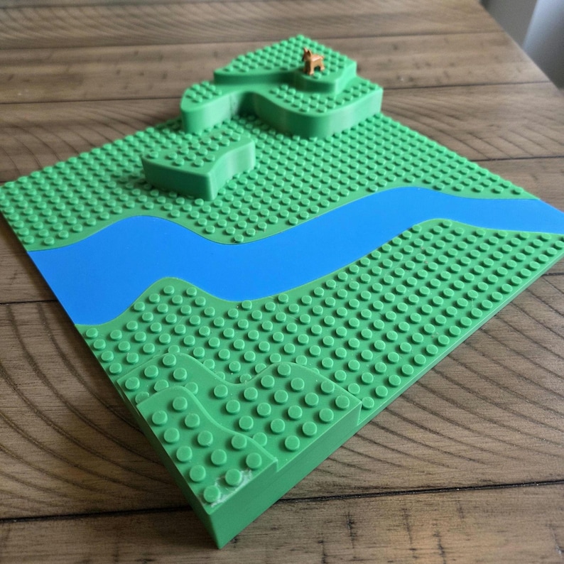 May include: A green building block baseplate with a blue river and raised green terrain. A small brown animal figurine is on the highest point. The baseplate is on a wooden surface.