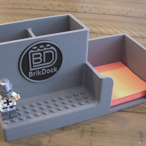 Op de afbeelding: Grijze bureau-organizer met vakken voor pennen en notitieblaadjes. Voorzien van een Lego-compatibele basis en een cirkelvormig logo met de tekst "BrikDock". Een Lego-figuur staat naast de organizer.