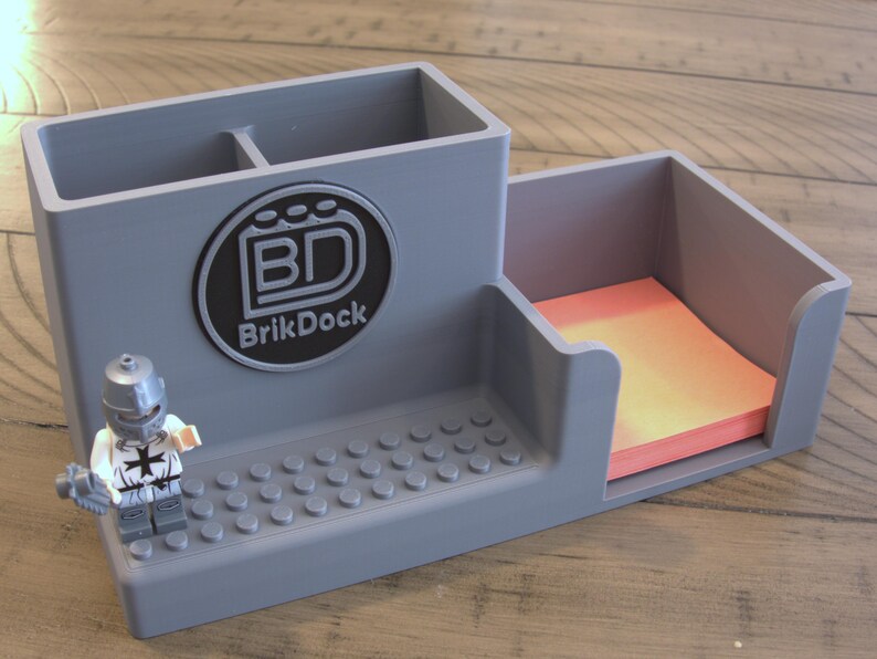 Op de afbeelding: Grijze bureau-organizer met vakken voor pennen en notitieblaadjes. Voorzien van een Lego-compatibele basis en een cirkelvormig logo met de tekst "BrikDock". Een Lego-figuur staat naast de organizer.