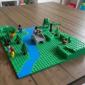 May include: A Lego diorama featuring a river, bridge, trees, and miniature figures. The base is green, with a blue river winding through the scene. Several Lego figures are positioned throughout the landscape, including a dog. The scene is set on a wooden table.