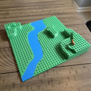 May include: A green building block landscape with a blue river winding through it. The landscape features raised areas and a small brown animal figure. The base is a square with a studded surface.