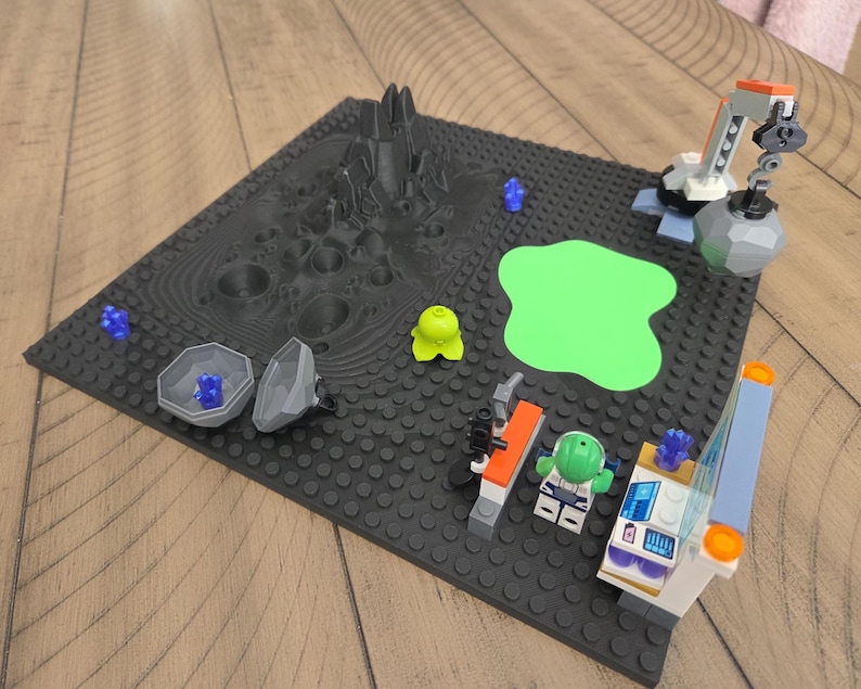 May include: A Lego space scene on a black baseplate. The scene includes a gray mountain, a green blob, a crane, and several Lego figures and accessories. The color palette is primarily gray, black, green, and blue.