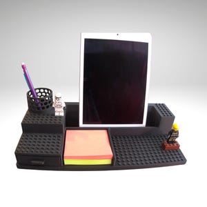 May include: Black desk organizer with a built-in tablet stand, pen holder, and drawer. Includes a stack of sticky notes and two toy figurines. The organizer is designed to resemble interlocking building blocks.