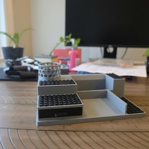 Pode incluir: Um organizador de secret&aacute;ria cinzento com um suporte cil&iacute;ndrico para canetas com recortes circulares, uma pequena gaveta com frente preta e v&aacute;rios n&iacute;veis planos cobertos com placas pretas com pinos. A estrutura &eacute; modular.