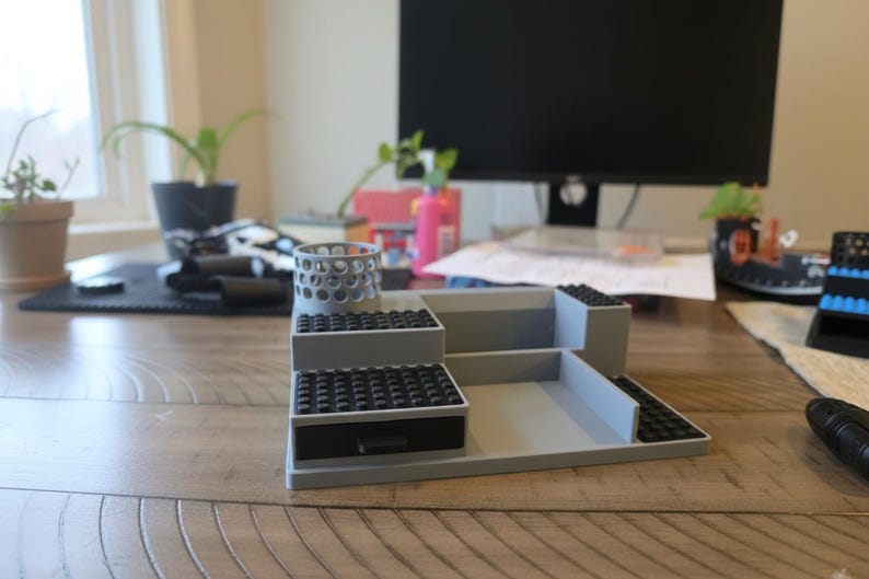 Pode incluir: Um organizador de secret&aacute;ria cinzento com um suporte cil&iacute;ndrico para canetas com recortes circulares, uma pequena gaveta com frente preta e v&aacute;rios n&iacute;veis planos cobertos com placas pretas com pinos. A estrutura &eacute; modular.