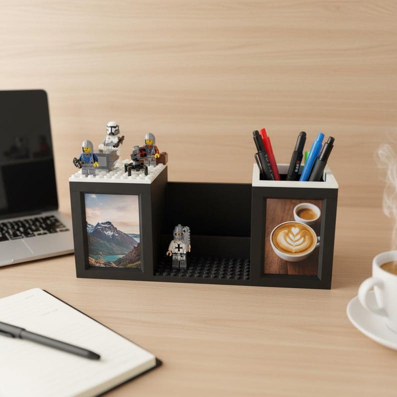May include: A black desk organizer with a white trim, featuring a photo frame, pen holders, and a central compartment. The organizer is decorated with Lego figures and a landscape photo. Pens and a coffee cup are also visible.