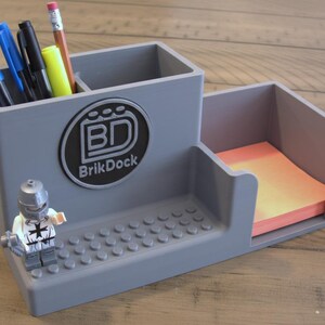 Op de afbeelding: Grijze bureau-organizer met een sectie voor pennen en potloden, een notitieblokhouder en een Lego-figuur. De organizer heeft het "BrikDock"-logo. Pennen, potloden en een notitieblok zijn zichtbaar.