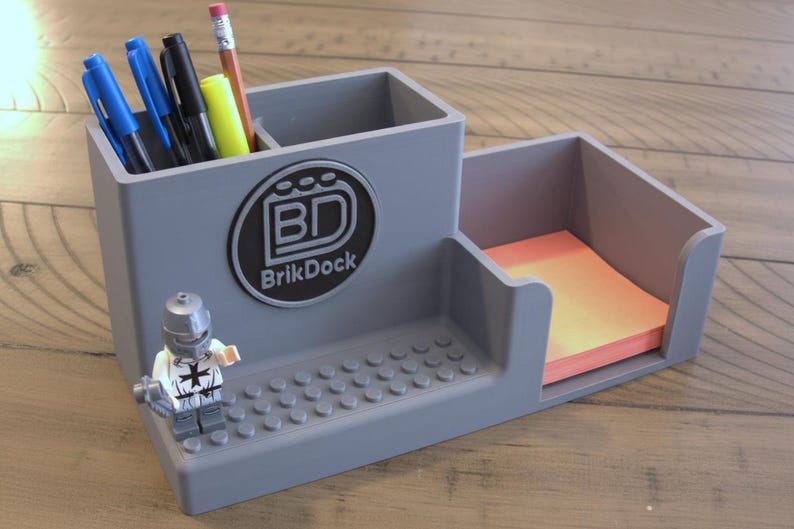 Op de afbeelding: Grijze bureau-organizer met een sectie voor pennen en potloden, een notitieblokhouder en een Lego-figuur. De organizer heeft het "BrikDock"-logo. Pennen, potloden en een notitieblok zijn zichtbaar.