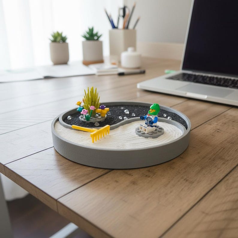 May include: A grey, circular miniature zen garden with white sand, black pebbles, and a yellow rake. The garden features Lego-style plants and a Lego figure. The garden sits on a wooden desk near a laptop.