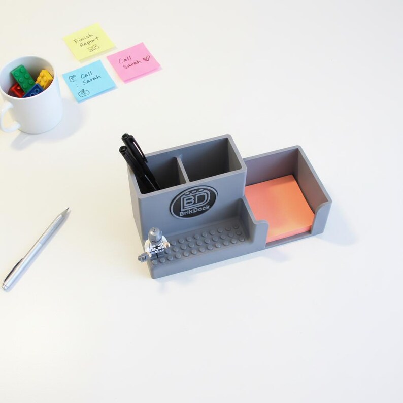 Op de afbeelding: Een grijze bureau-organizer met een ingebouwde notitieblokhouder en pennenvakken. De organizer heeft de tekst "BrikDock" op de voorkant en een klein Lego-figuur. Een witte mok met kleurrijke blokken, een zilveren pen en plaknotities zijn ook zichtbaar.