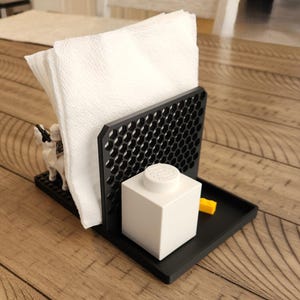 May include: A black napkin holder with a honeycomb design, holding white napkins. A white Lego block and a yellow Lego brick are on the tray. A small toy figure is on the base.