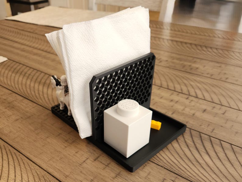 May include: A black napkin holder with a honeycomb design, holding white napkins. A white Lego block and a yellow Lego brick are on the tray. A small toy figure is on the base.