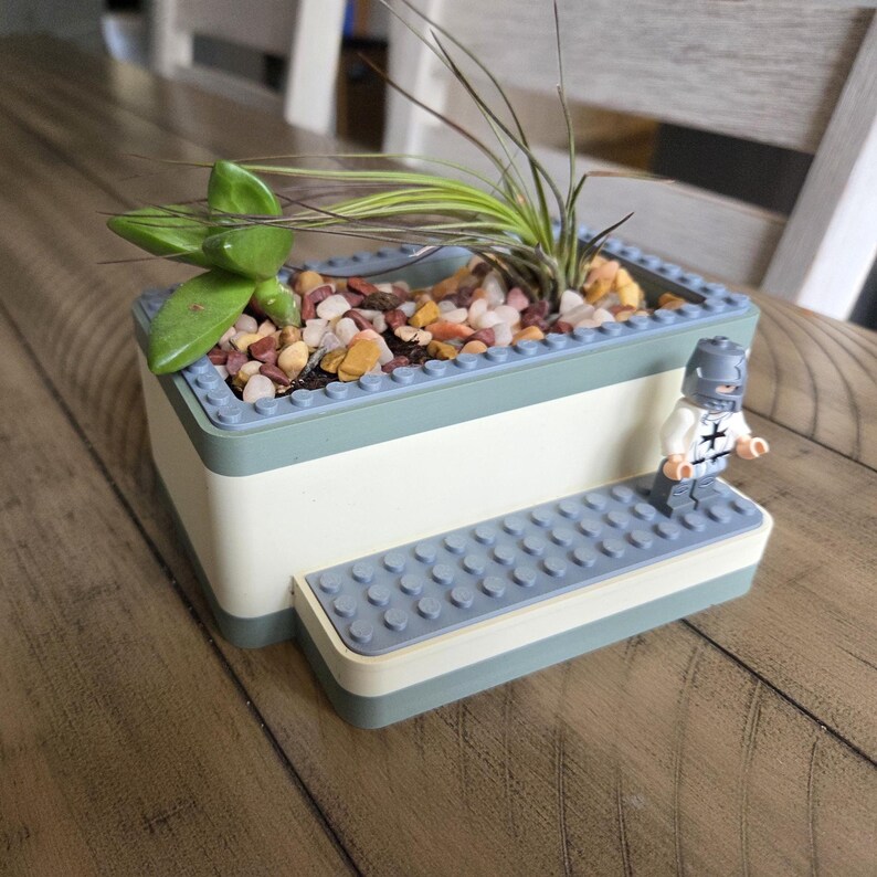 May include: A rectangular planter with a light blue and white design, filled with small colorful stones and greenery. A small Lego figure of a knight stands on a step attached to the planter.