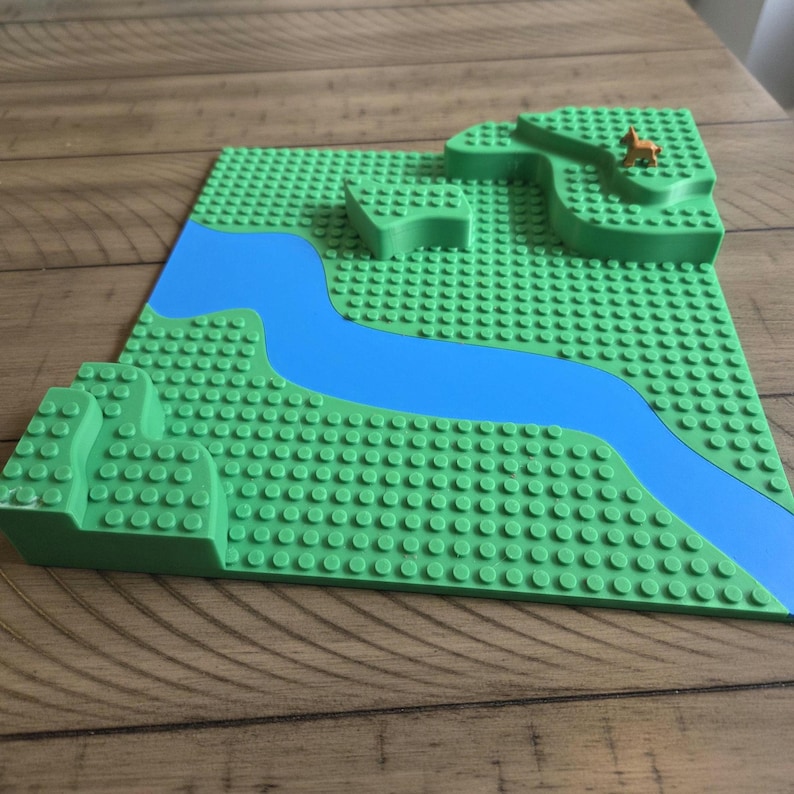 May include: A green building block baseplate with a blue river design and raised green terrain. A small brown animal figurine is on the raised terrain. The baseplate is on a wooden surface.
