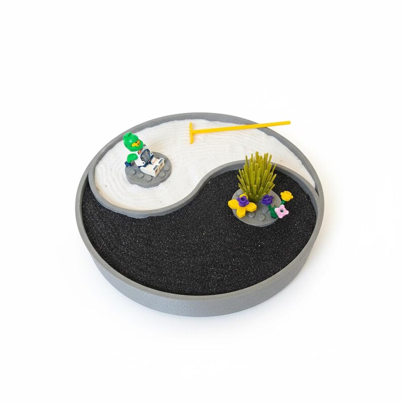 May include: A miniature Zen garden in a Yin Yang design. The garden features white sand, black sand, a yellow rake, and Lego figures. The garden is in a grey circular tray with a white background.