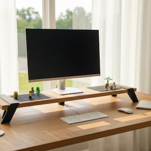 Puede incluir: Un escritorio de madera con un monitor de computadora, teclado y ratón. Un soporte de monitor de madera con patas negras sostiene el monitor. Pequeñas plantas decorativas y figuritas están en el soporte. Una computadora portátil y un cargador inalámbrico también están en el escritorio.