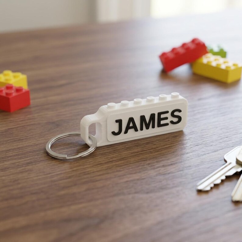 May include: A white rectangular plastic keychain featuring a top row of small circular studs and the name JAMES printed in bold black capital letters on the side, attached to a circular silver metal split ring for holding keys securely.