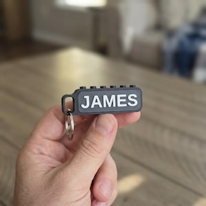 May include: A black rectangular keychain featuring the name JAMES in raised white capital letters. The top edge of the keychain has six small circular studs, and a silver metal split ring is attached to the left side of the black frame.