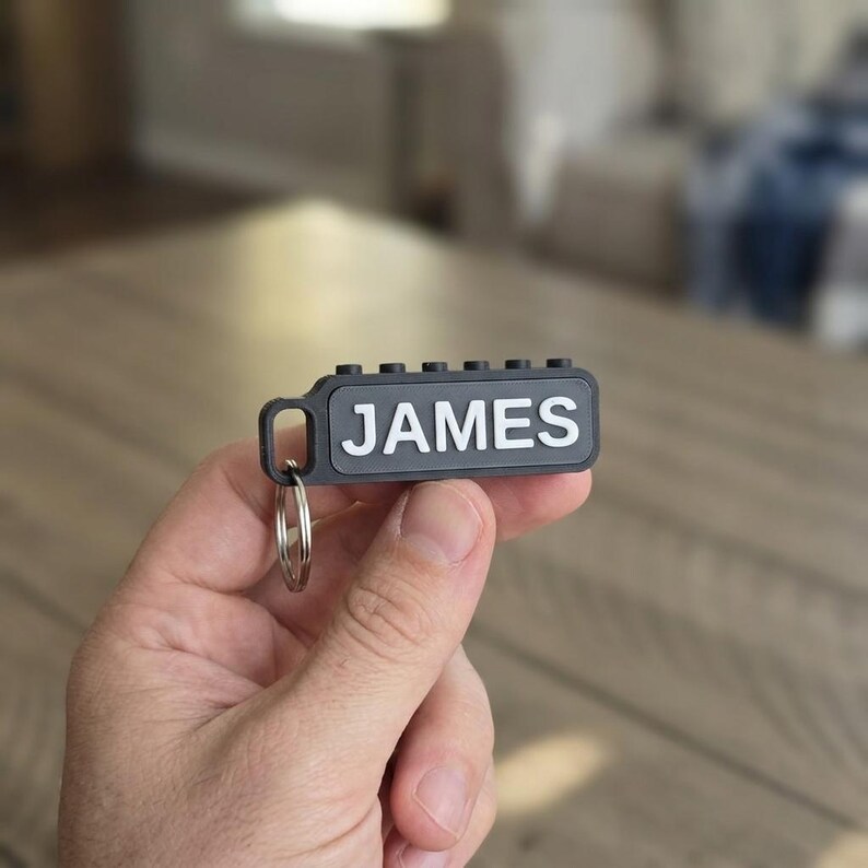 May include: A black rectangular keychain featuring the name JAMES in raised white capital letters. The top edge of the keychain has six small circular studs, and a silver metal split ring is attached to the left side of the black frame.