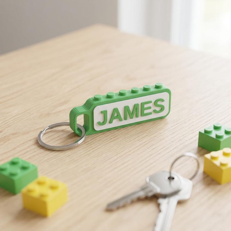 May include: A green and white rectangular keychain featuring the name JAMES in raised lettering, designed with a block-like texture on top and a metal ring attached to one end, accompanied by small green and yellow plastic building blocks.