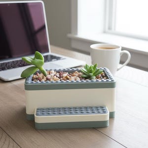 May include: A rectangular, two-toned planter with a small succulent plant. The planter is white with a gray trim and contains small pebbles. A matching small rectangular piece sits in front of the planter. A laptop and a mug of coffee are in the background.