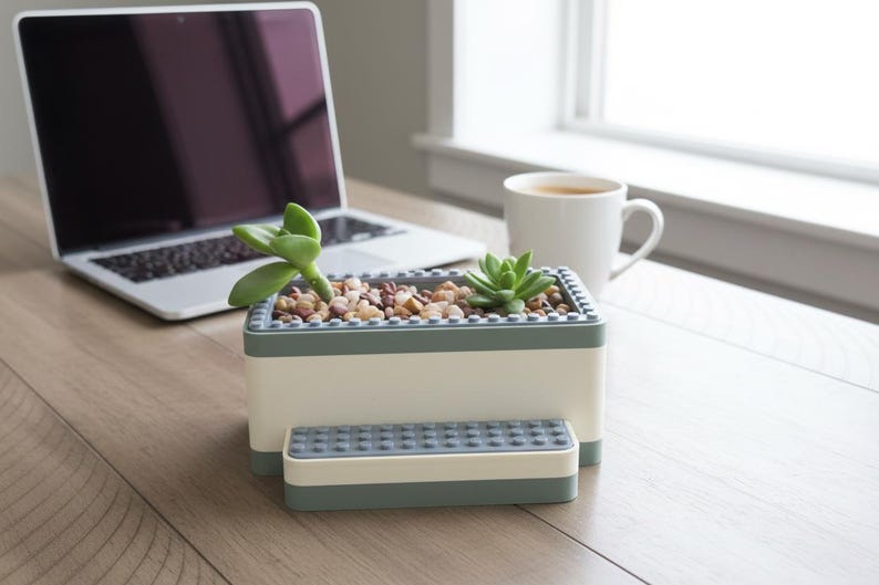 May include: A rectangular, two-toned planter with a small succulent plant. The planter is white with a gray trim and contains small pebbles. A matching small rectangular piece sits in front of the planter. A laptop and a mug of coffee are in the background.