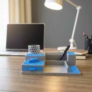 Pode incluir: Um organizador de secret&aacute;ria cinzento com design em camadas e placas de blocos de constru&ccedil;&atilde;o azuis nas superf&iacute;cies. Inclui uma pequena gaveta azul com puxador, um porta-canetas cil&iacute;ndrico com padr&atilde;o perfurado e compartimentos abertos.