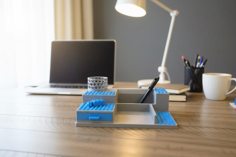 Pode incluir: Um organizador de secret&aacute;ria cinzento com design em camadas e placas de blocos de constru&ccedil;&atilde;o azuis nas superf&iacute;cies. Inclui uma pequena gaveta azul com puxador, um porta-canetas cil&iacute;ndrico com padr&atilde;o perfurado e compartimentos abertos.