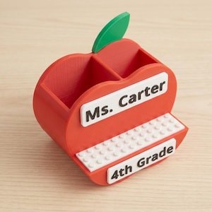 May include: A red apple-shaped desk organizer with a green leaf, featuring two compartments. It has a white rectangular plaque with the black text Ms. Carter, a white studded base, and a white plaque with the black text 4th Grade on the front.