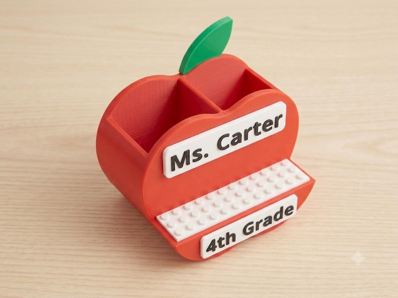 May include: A red apple-shaped desk organizer with a green leaf, featuring two compartments. It has a white rectangular plaque with the black text Ms. Carter, a white studded base, and a white plaque with the black text 4th Grade on the front.