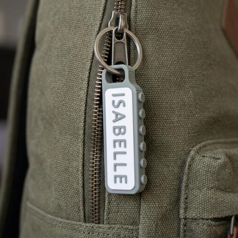 May include: A gray rectangular keychain with a white center panel featuring the name ISABELLE in raised gray letters. The keychain has a side edge with small circular protrusions and is attached to a silver metal split ring on a green canvas bag.