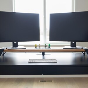Pode incluir: Uma secret&aacute;ria preta com dois grandes monitores de computador, um teclado e um suporte de monitor de madeira. O suporte tem pernas pretas e cont&eacute;m pequenas figuras de Lego. A secret&aacute;ria est&aacute; em frente a uma janela.