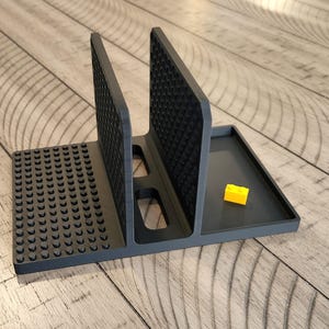 May include: A dark grey plastic organiser with several compartments. It features two vertical dividers with textured surfaces, a base with circular studs, and a small tray. A yellow building block is in the tray.