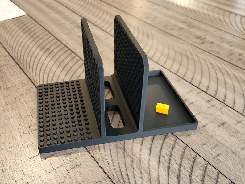 May include: A dark grey plastic organiser with several compartments. It features two vertical dividers with textured surfaces, a base with circular studs, and a small tray. A yellow building block is in the tray.