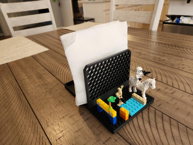 May include: Black napkin holder with a geometric pattern, holding white napkins. The base is decorated with Lego figures, including a Stormtrooper on a white horse. A fun home decor item.