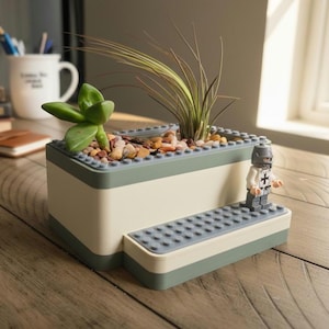 May include: A two-tone planter with a light green and off-white design, filled with small rocks and greenery. A Lego figurine of a knight stands on a small platform attached to the planter. The planter is on a wooden surface.