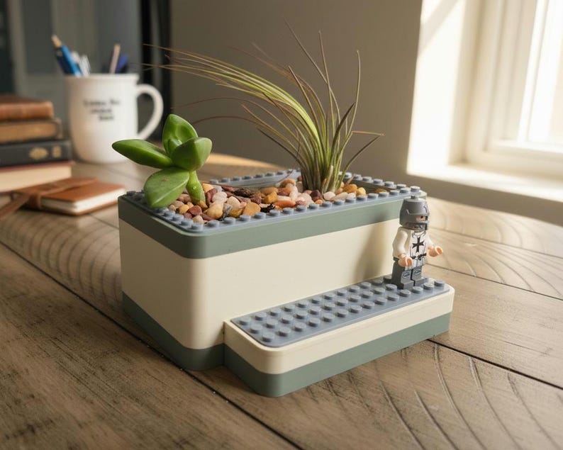 May include: A two-tone planter with a light green and off-white design, filled with small rocks and greenery. A Lego figurine of a knight stands on a small platform attached to the planter. The planter is on a wooden surface.