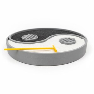 May include: A grey Yin Yang zen garden with black and white sand, two grey stone-like accents, and a yellow rake. The garden is a symbol of balance and harmony, ideal for a desk or tabletop.